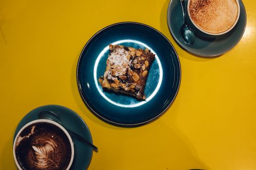 Free A top-down view of a brownie on a blue plate with cappuccinos and cocoa drinks on a vibrant yellow background. Stock Photo
