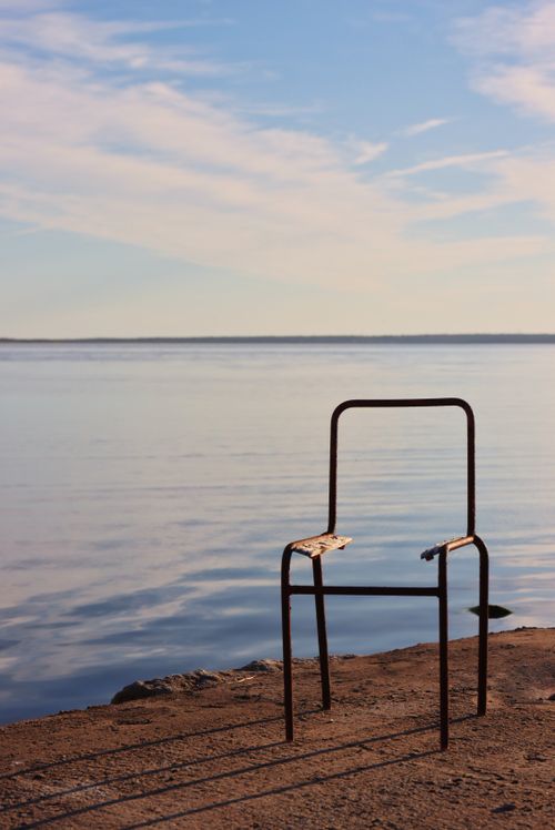 Free A weathered chair on a serene lakeshore under a vibrant sunset sky. Stock Photo