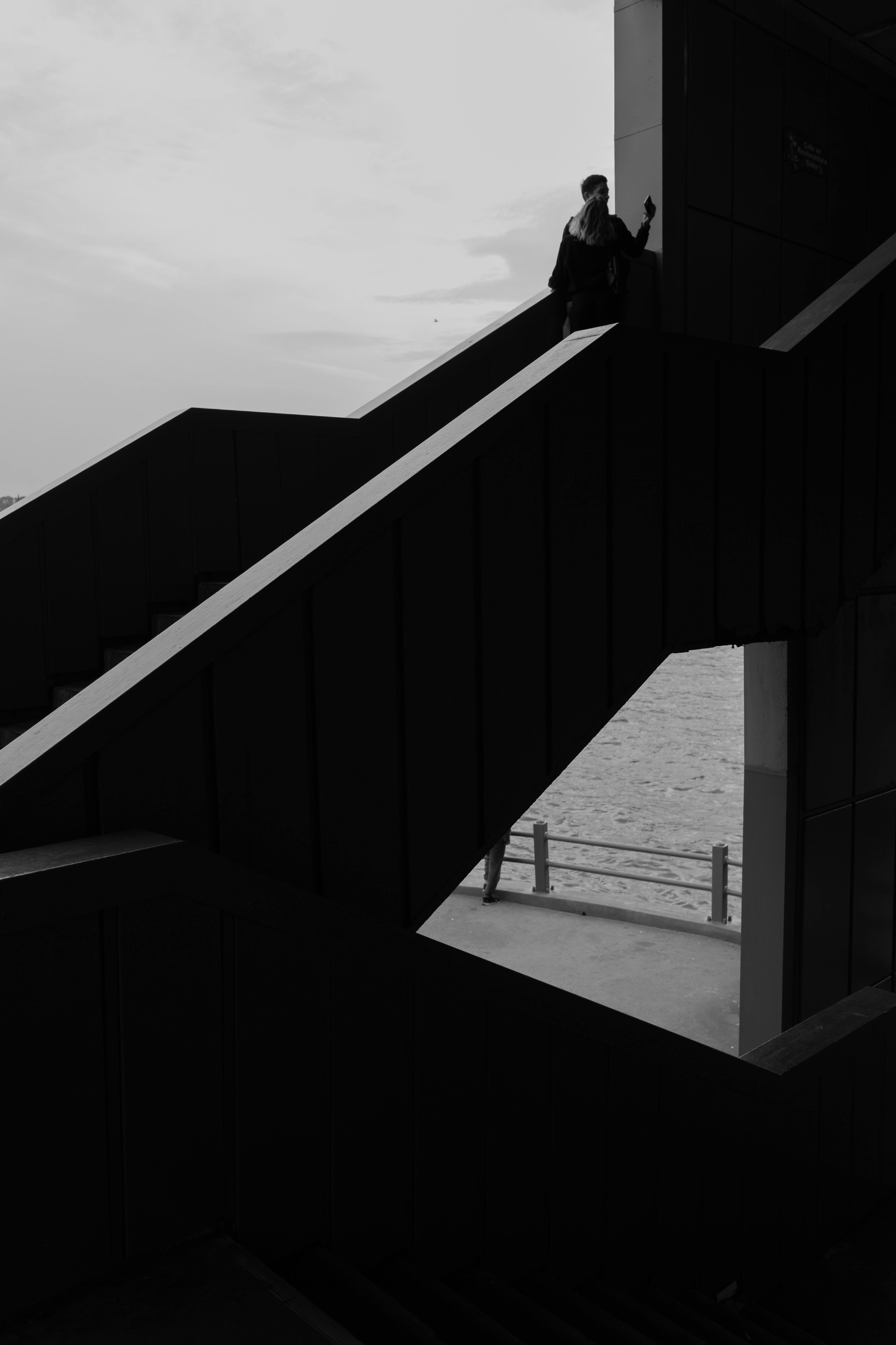 Ücretsiz Deniz kenarında geometrik bir merdiven üzerinde duran bir kişinin dramatik siyah beyaz silüeti. Stok Fotoğraflar