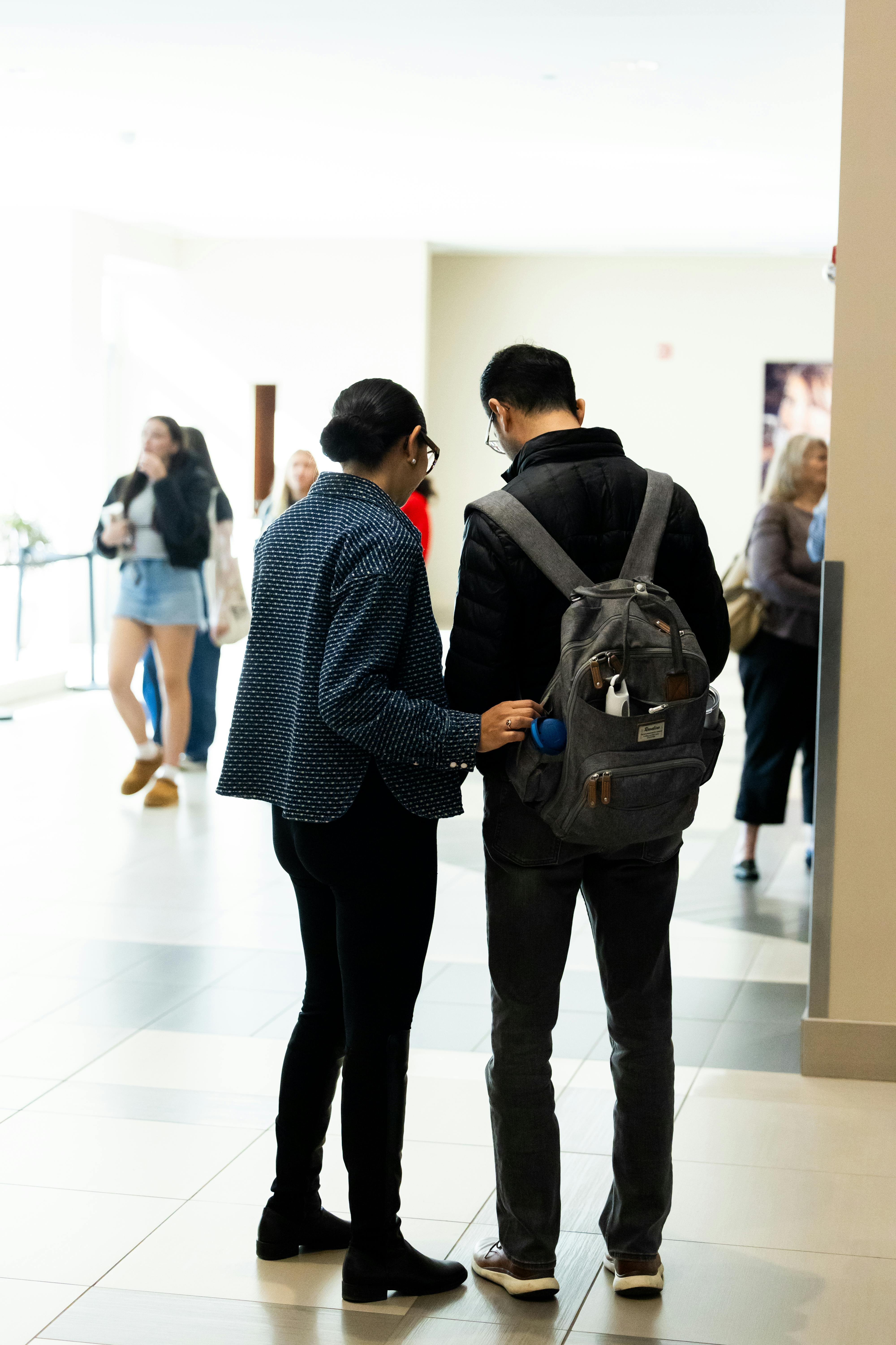 grátis Dois adultos conversando informalmente em um corredor interno moderno, com outras pessoas ao fundo. Foto profissional