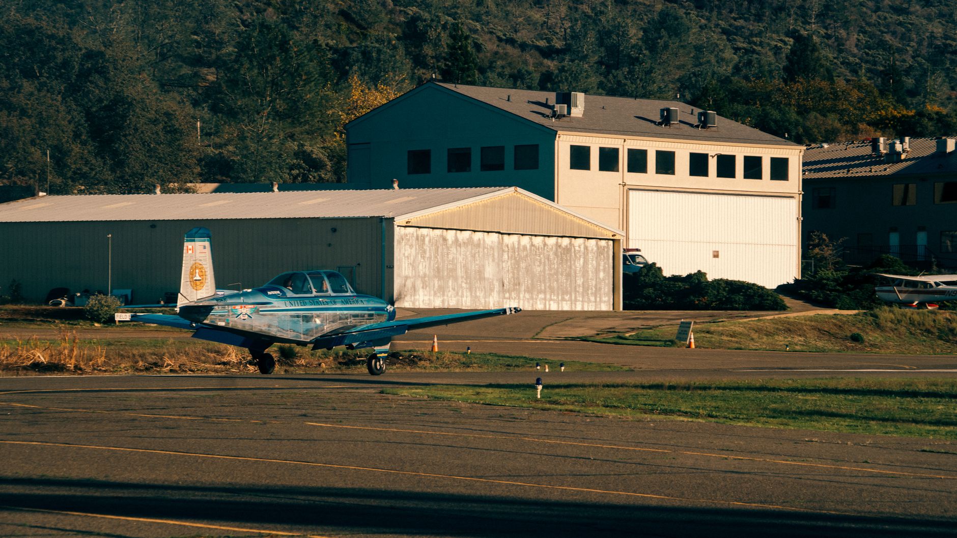 A blue small aircraft parked on an airfield runway near a hangar during daytime.