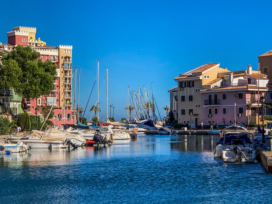 Vibrant marina scene with yachts docked along colorful Mediterranean-style buildings under a clear blue sky.