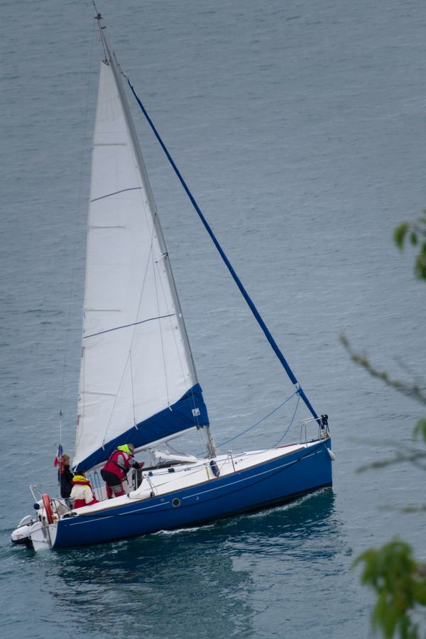 Voilier à coque bleue et équipage en vestes rouges naviguant sur une mer grise et calme