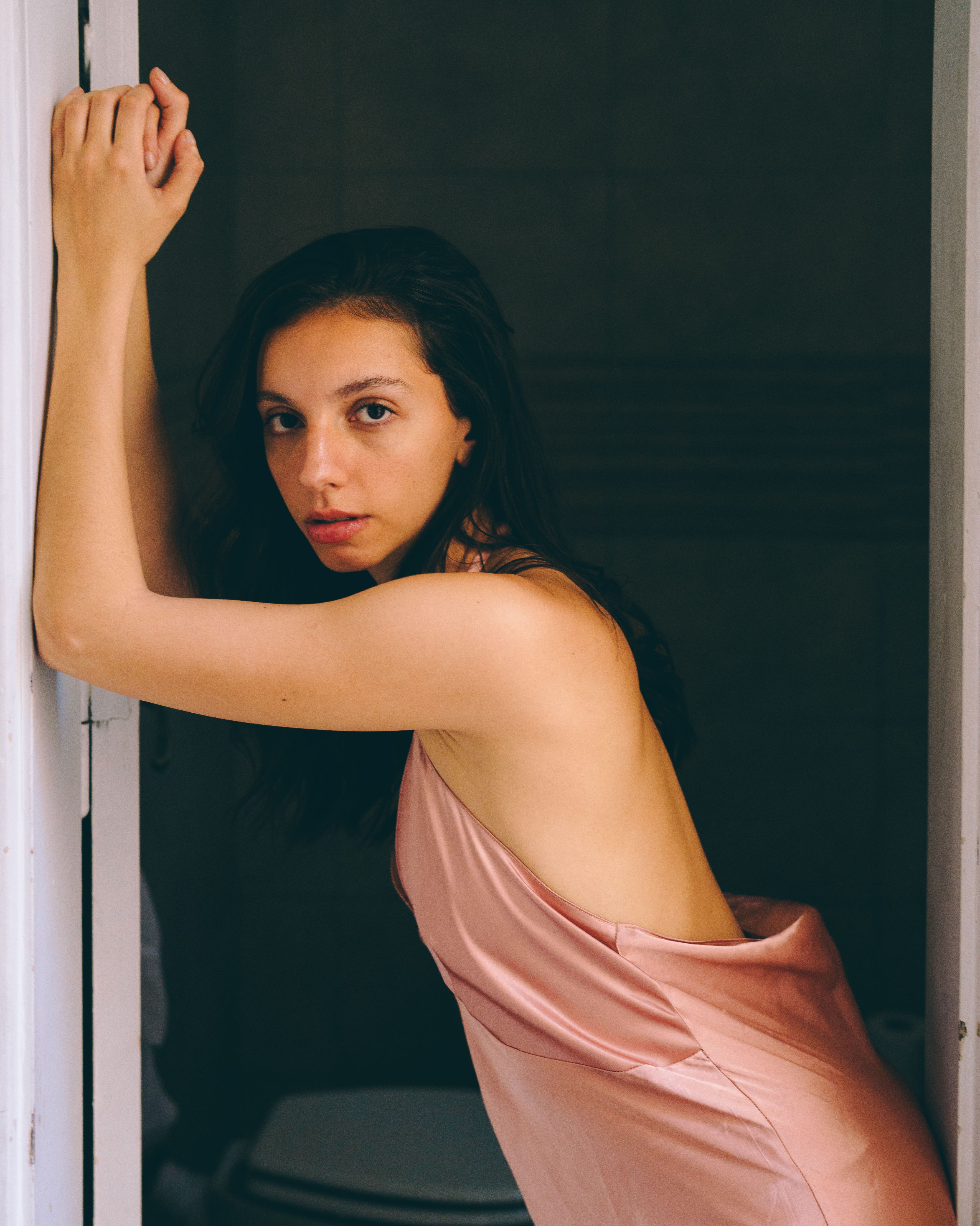 A stylish adult woman poses in a pink dress, leaning against a doorway in an indoor setting.