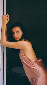 A woman in a pink silky dress gracefully poses indoors, exuding elegance.