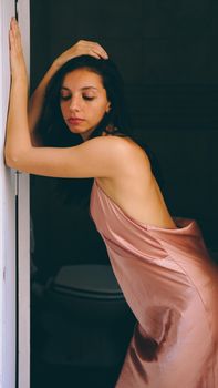 Woman in a soft pink satin dress leaning on a door frame, captured indoors.