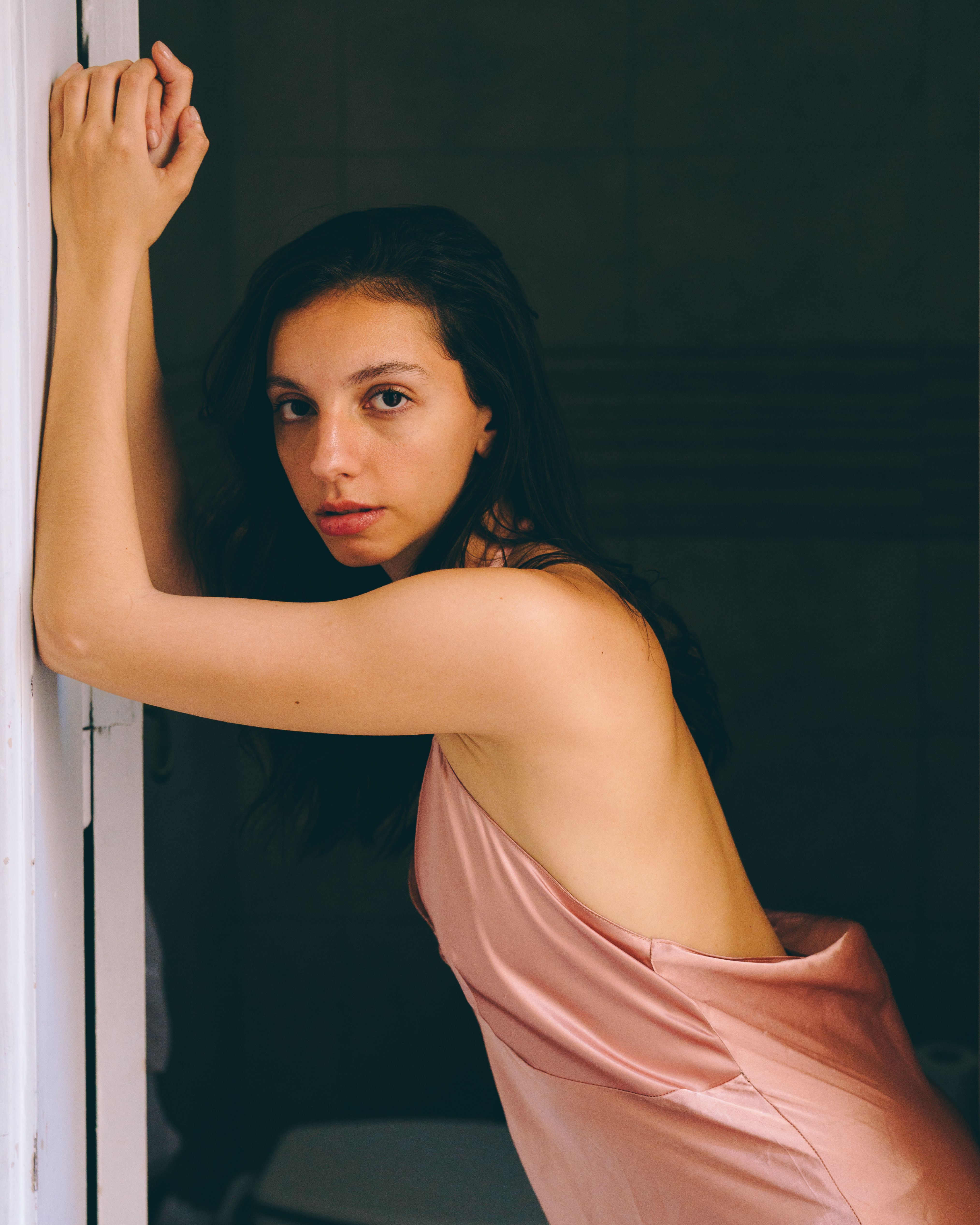 Woman leaning on doorframe in soft lighting, wearing a silk dress.