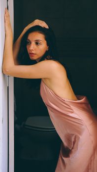 Elegant woman in a pink silk dress posing indoors by a doorway.