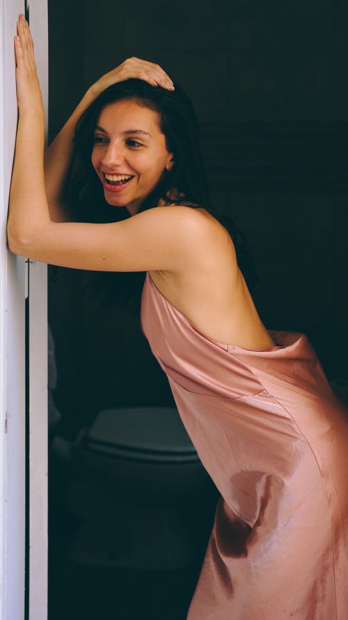 Free A joyful adult woman in a pink silk dress poses playfully by an indoor doorway. Stock Photo