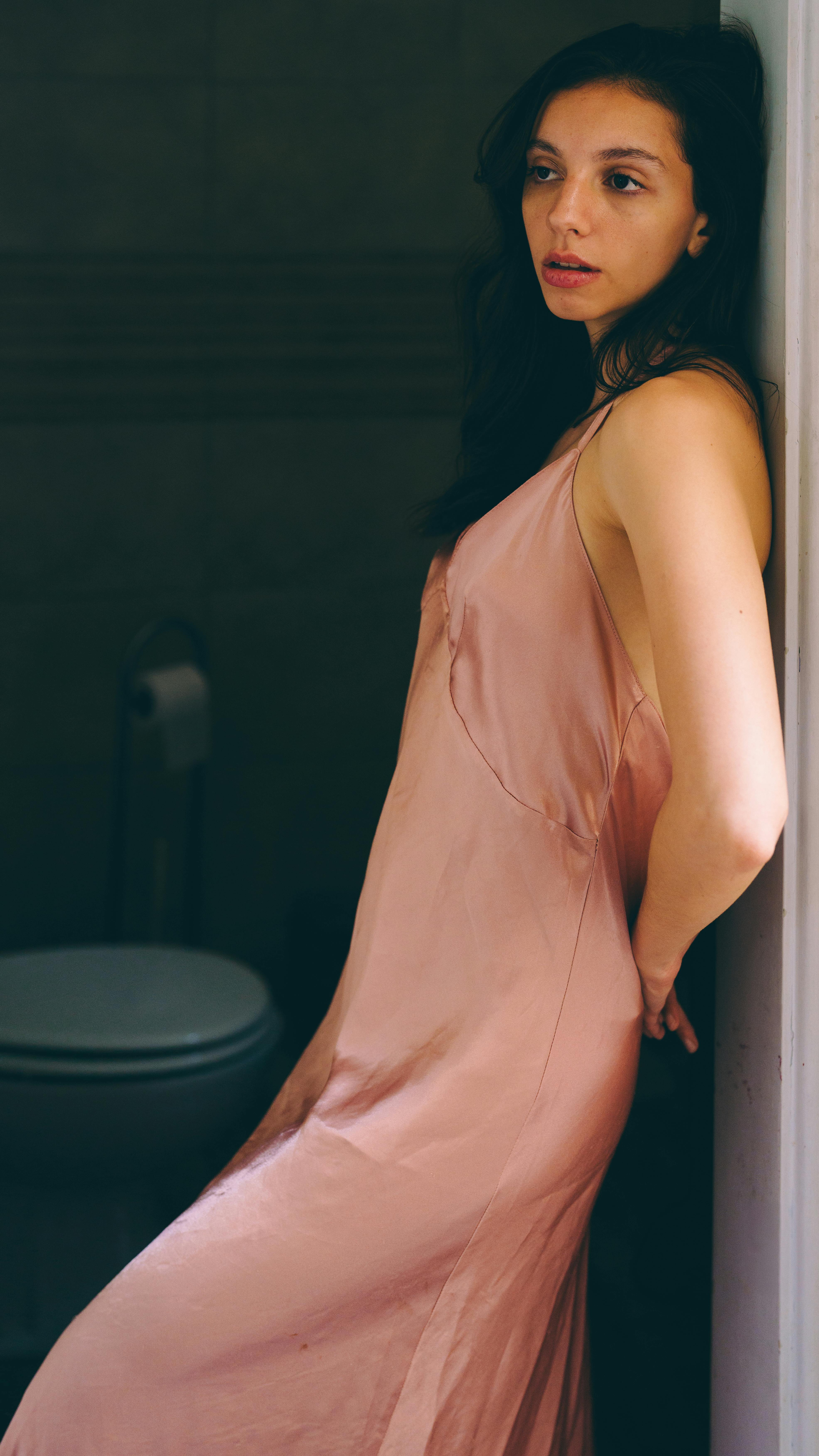 A woman in a pink dress leaning against a wall, deep in thought indoors.