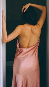 A woman in a backless pink silk dress posing gracefully indoors.
