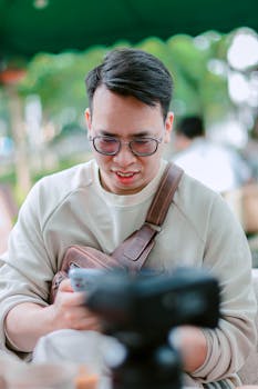 A smiling young man wearing glasses interacts with his smartphone outdoors.