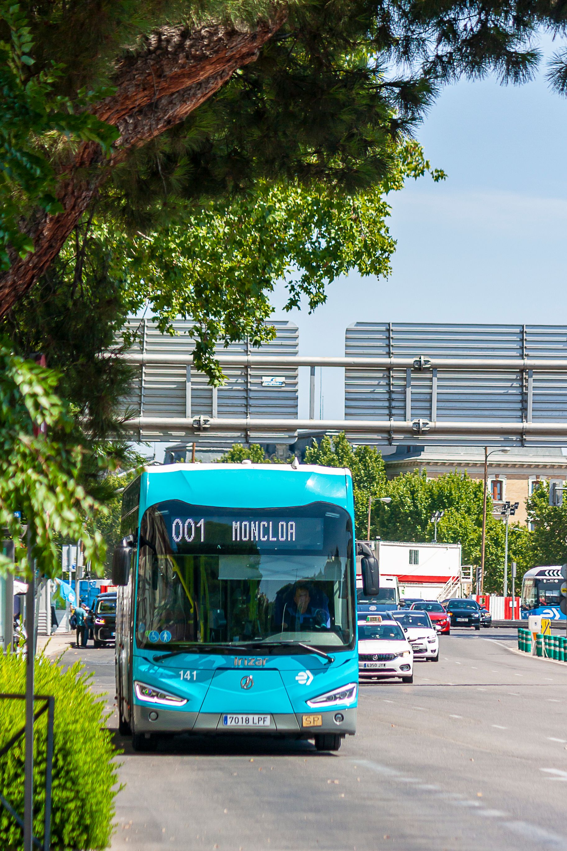 A city bus, number 001, in Madrid heading towards Moncloa on a sunny day, surrounded by lush greenery.