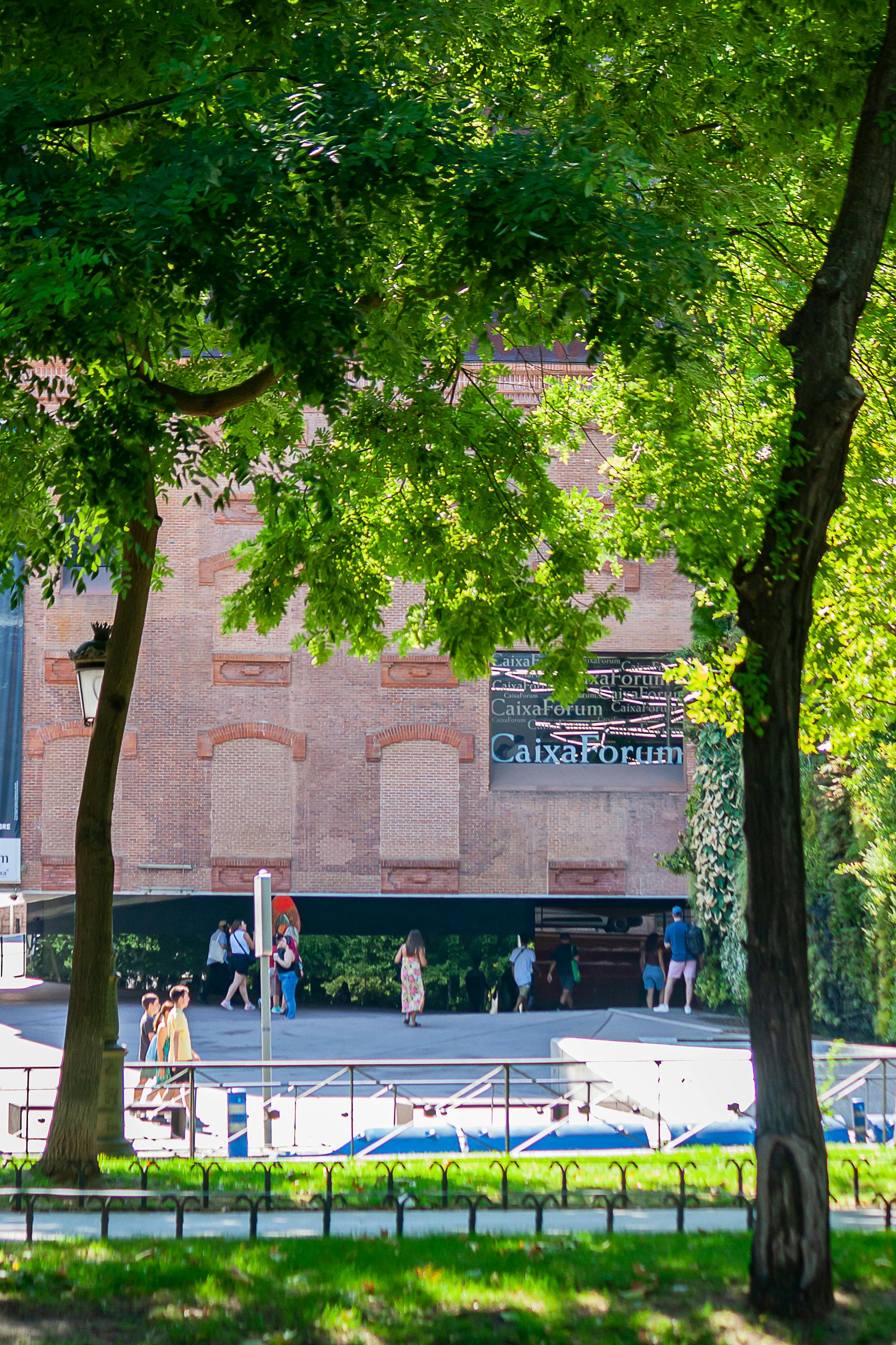 CaixaForum Madrid facade with people walking and lush green trees on a sunny day.