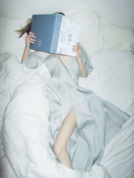 An adult woman in a silky gown reads in bed, hiding her face with a book.