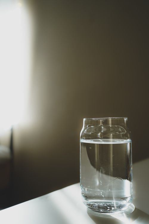 Free A single glass of water sits on a table in a softly lit room, creating a minimalist aesthetic. Stock Photo