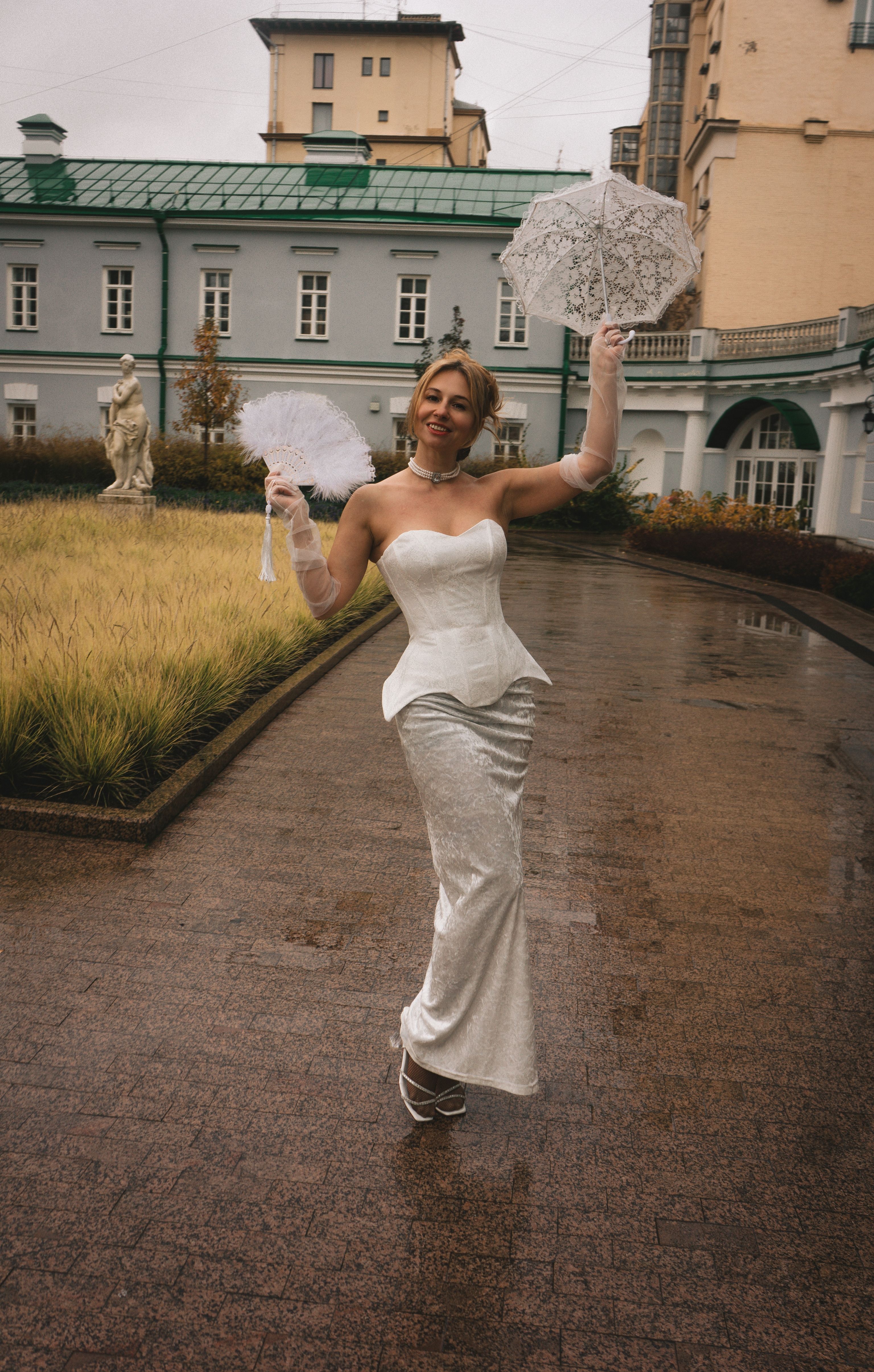 An elegant woman in a white gown holds a lace parasol, standing outdoors on a rainy day.