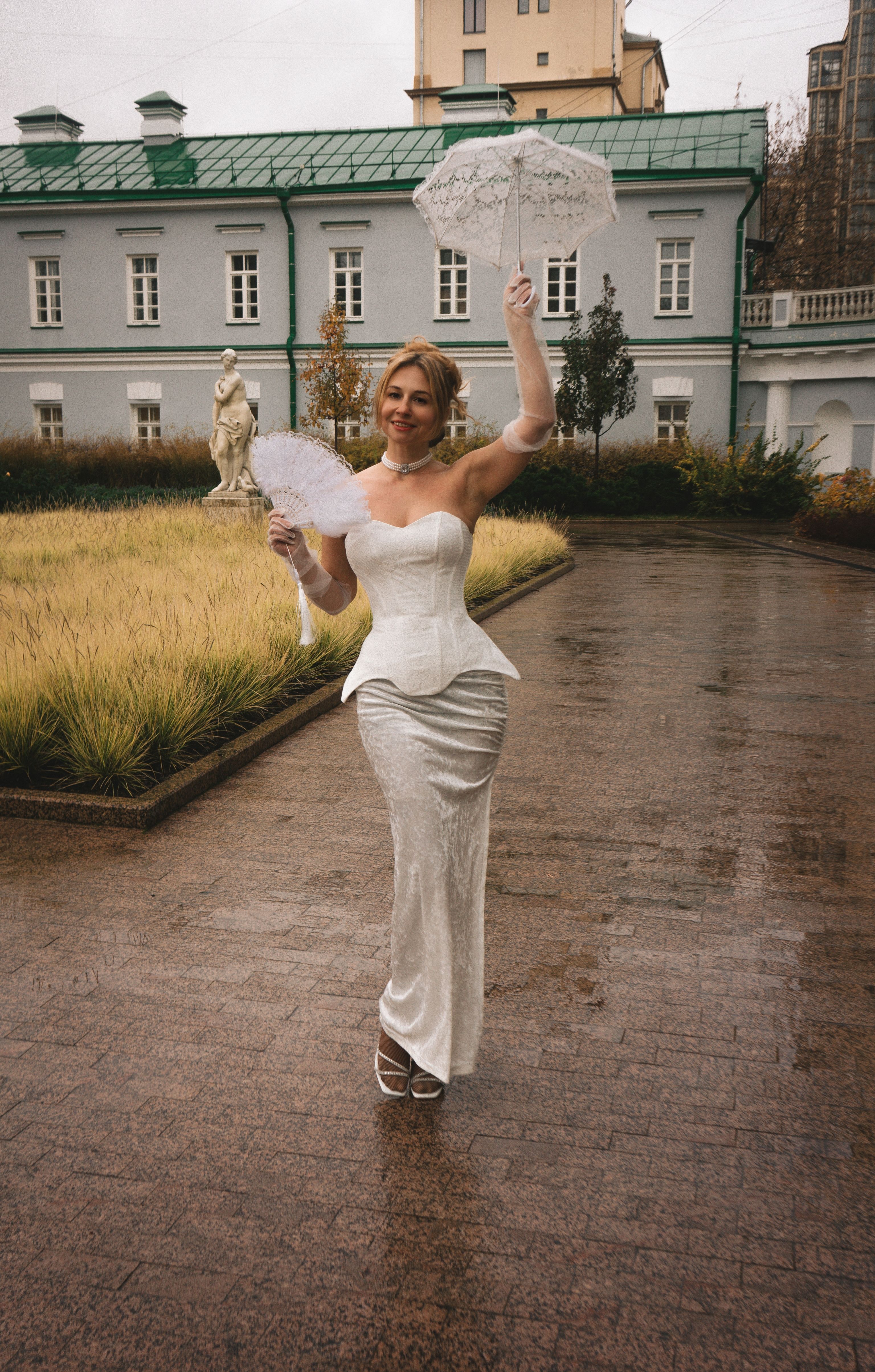 A woman in an elegant white dress poses with umbrellas in a garden setting.