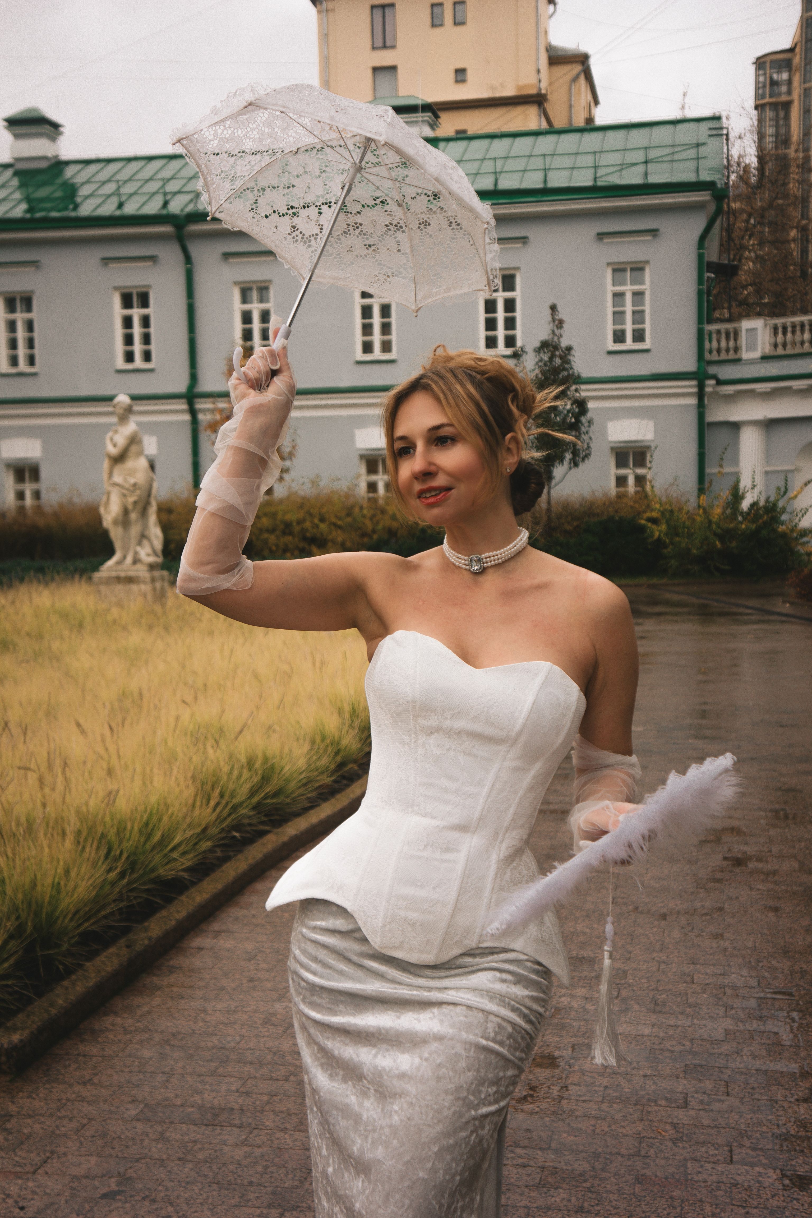 Elegant woman in white bridal attire holding a lace umbrella outdoors near a classic building.