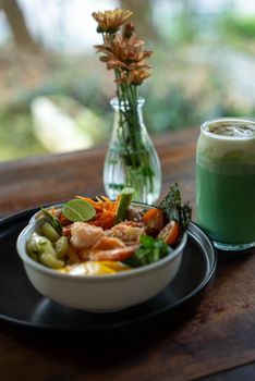 Fresh salad bowl with shrimp and vegetables beside a refreshing green smoothie and flowers on a wooden table.