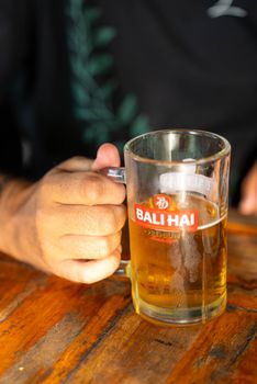 A hand holding a frosty glass of Bali Hai beer on a wooden table, showcasing branding.