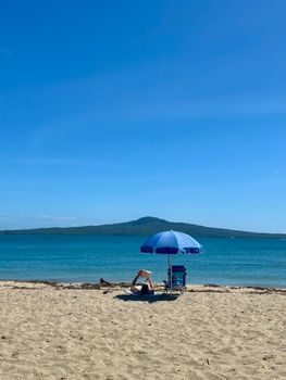 Person enjoying beach reading under a blue umbrella with Rangitoto Island view in Auckland, New Zealand.