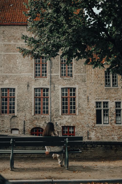 Free Woman sitting alone on a park bench near a historic brick building with tree overhead. Stock Photo