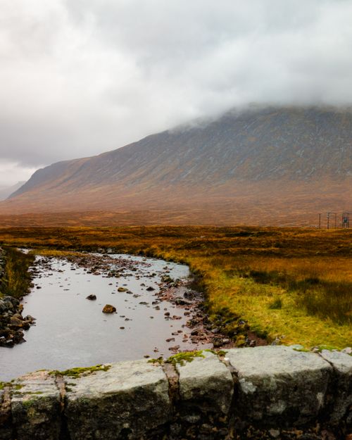 Free A serene view of Glencoe, Scotland featuring a mountain, stream, and cloudy sky. Stock Photo