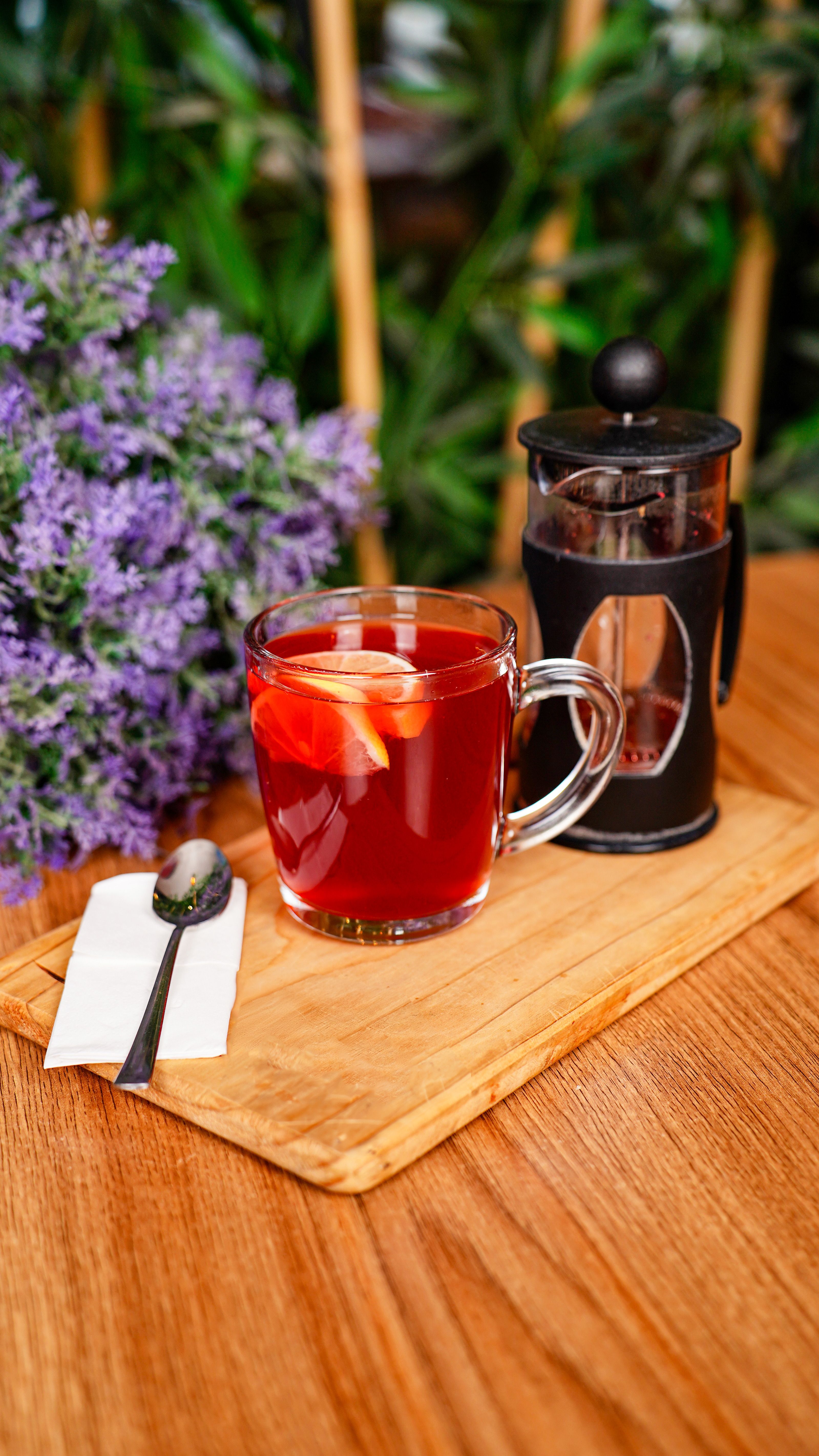 A warm cup of herbal tea with lemon slices beside a French press on a wooden tray, surrounded by lavender flowers.