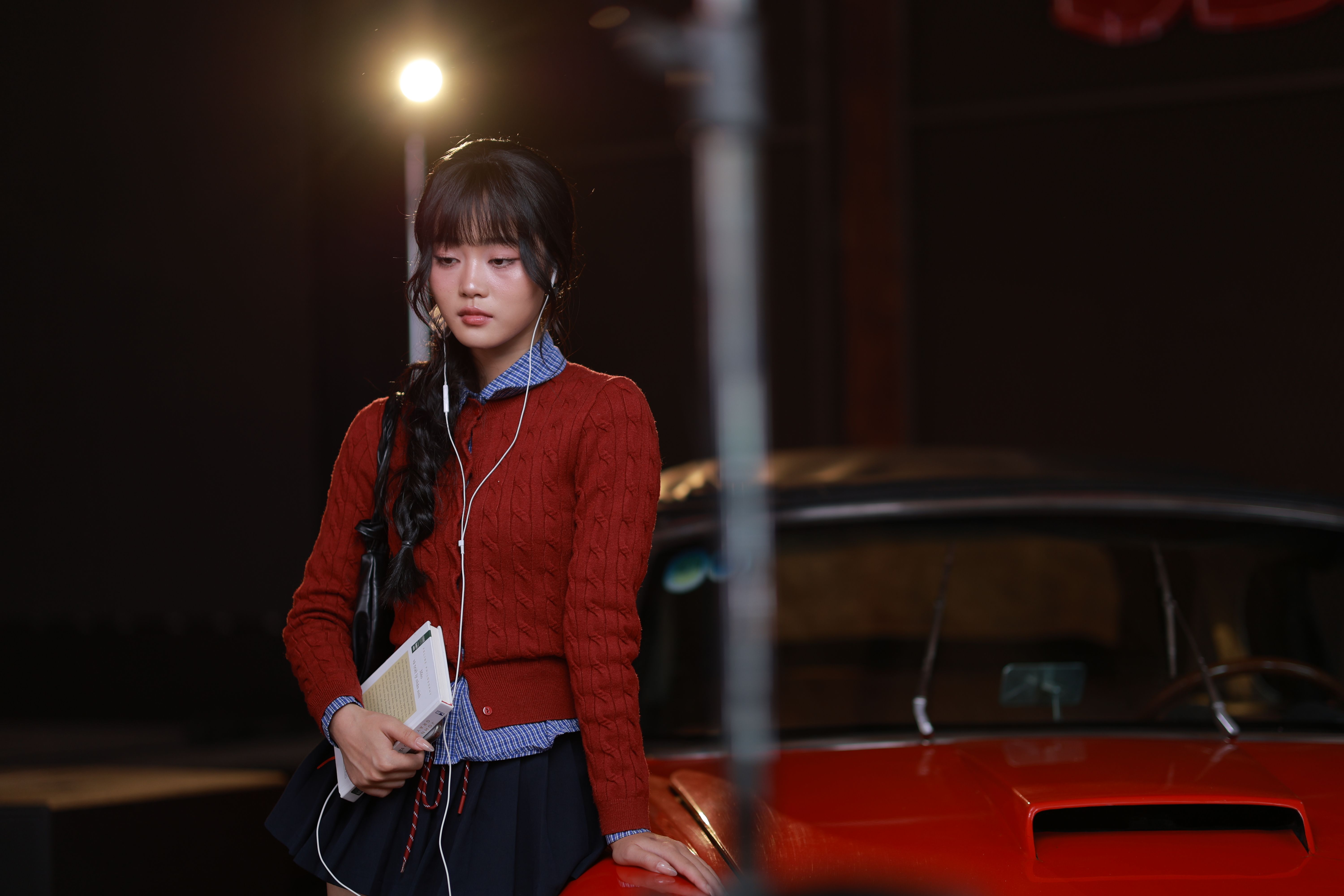 A young woman with earphones and a book stands indoors by a classic car, lit dramatically.