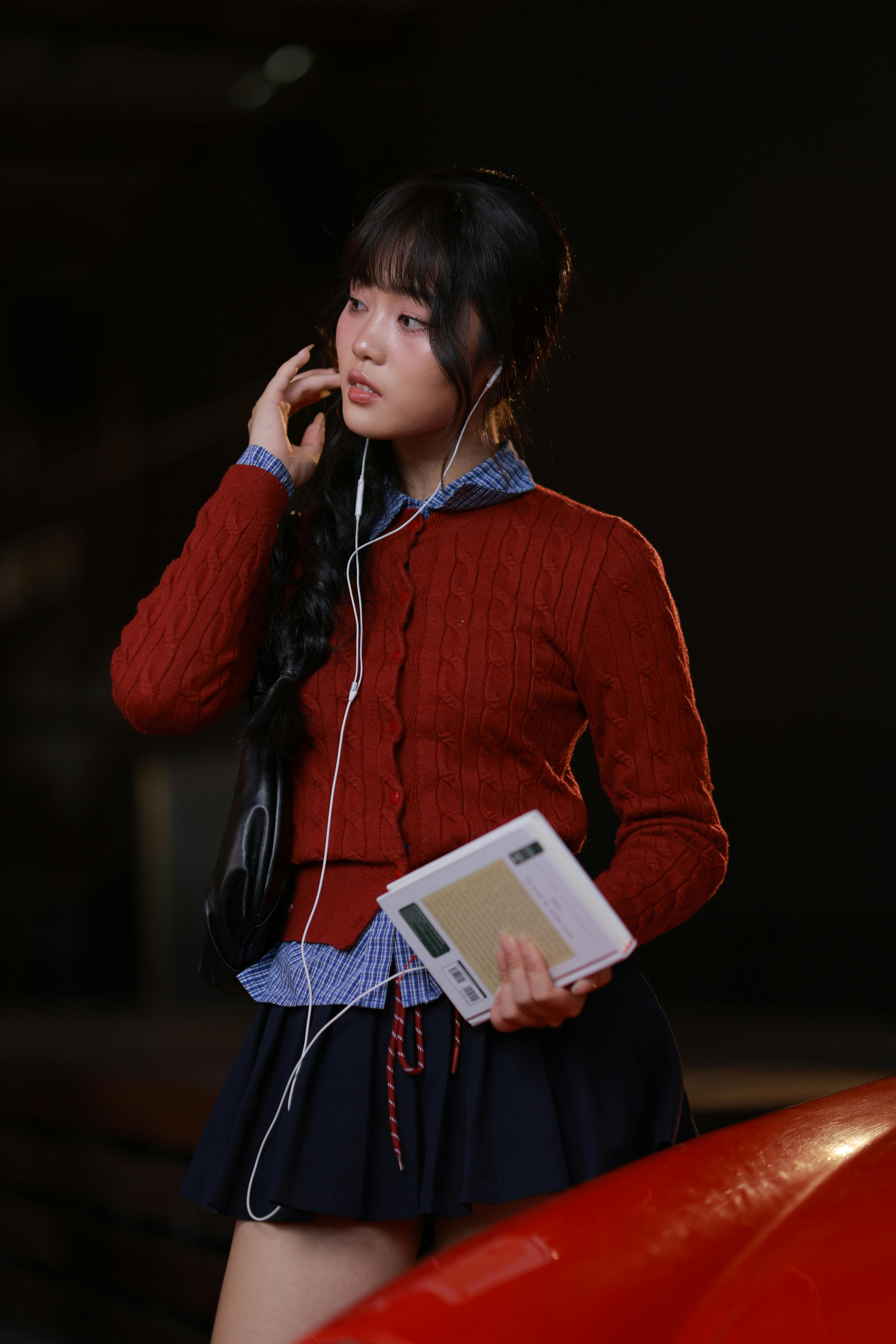 grátis Uma jovem ouve música com fones de ouvido enquanto lê um livro à noite, vestindo uma roupa elegante. Foto profissional