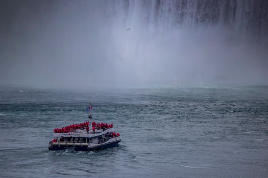 Tour boat approaches the majestic mist of Niagara Falls with passengers wearing red raincoats.