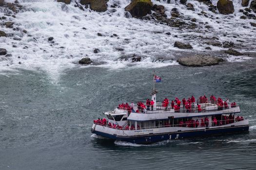 Tourist boat in red raincoats cruising near Niagara Falls rapids. Exciting adventure.