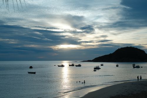 Free Tranquil beach scene at sunset with boats and silhouettes on a calm sea. Stock Photo