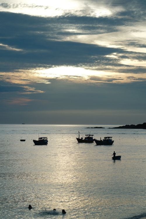 Free Boats rest peacefully on calm waters under a dramatic evening sky with people enjoying the sea. Stock Photo
