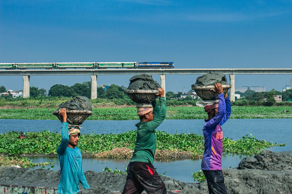 Three workers carry baskets of clay on their heads beside a river with a train on a bridge in the background.