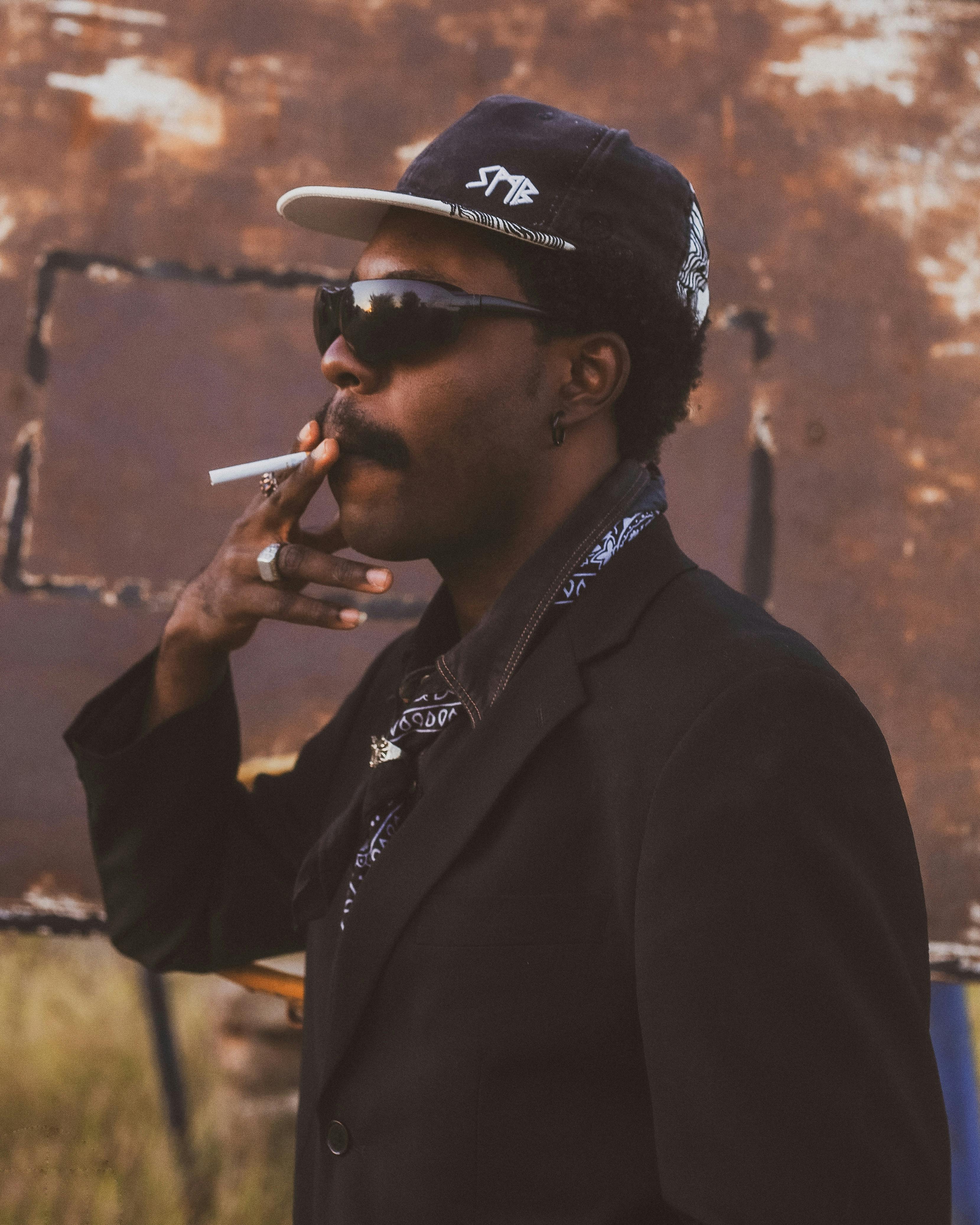 A man dressed in black smokes a cigarette outdoors. Fashionable look with sunglasses and cap.