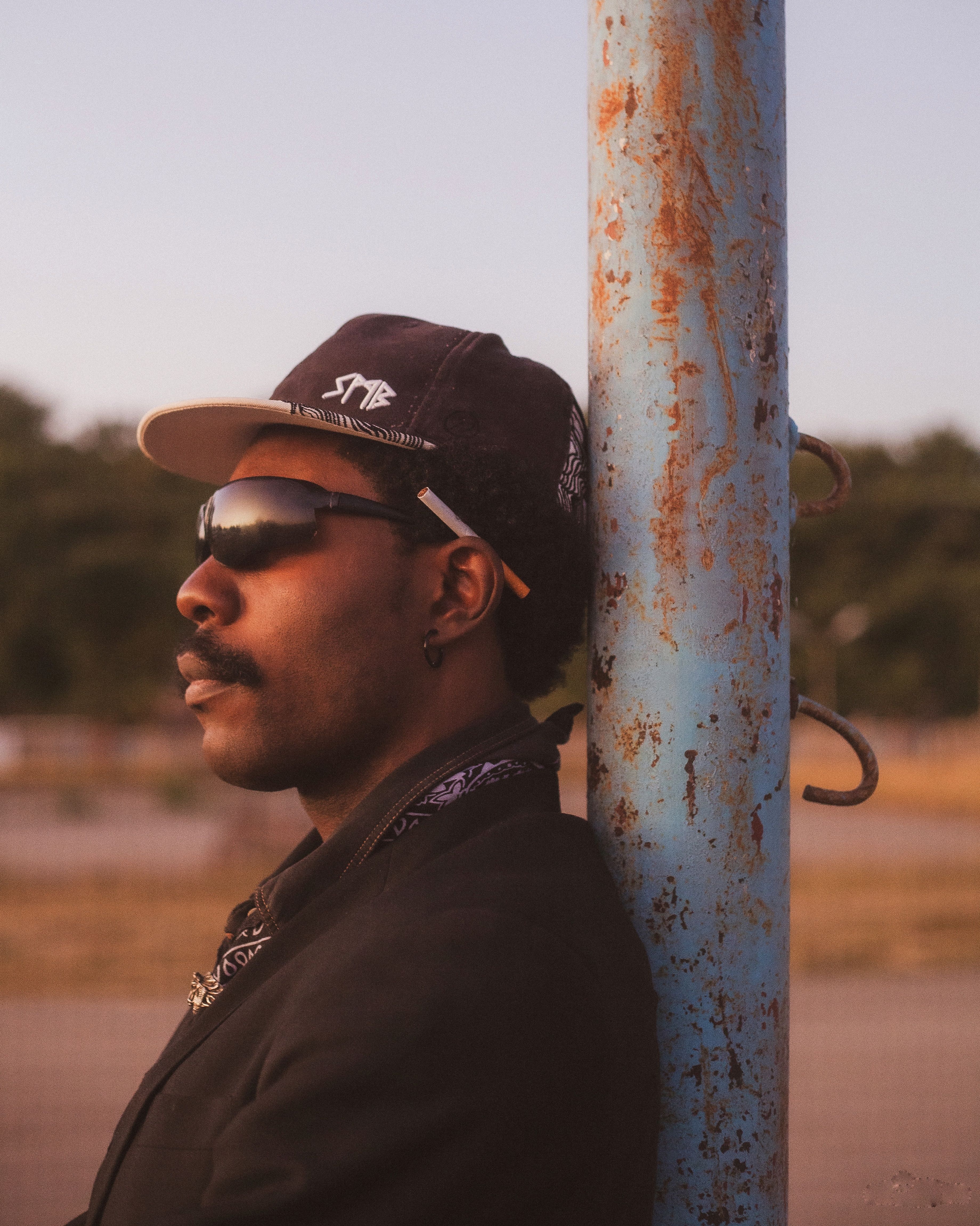 Stylish man in sunglasses and hat stands at dusk, leaning on a rusty pole outdoors.