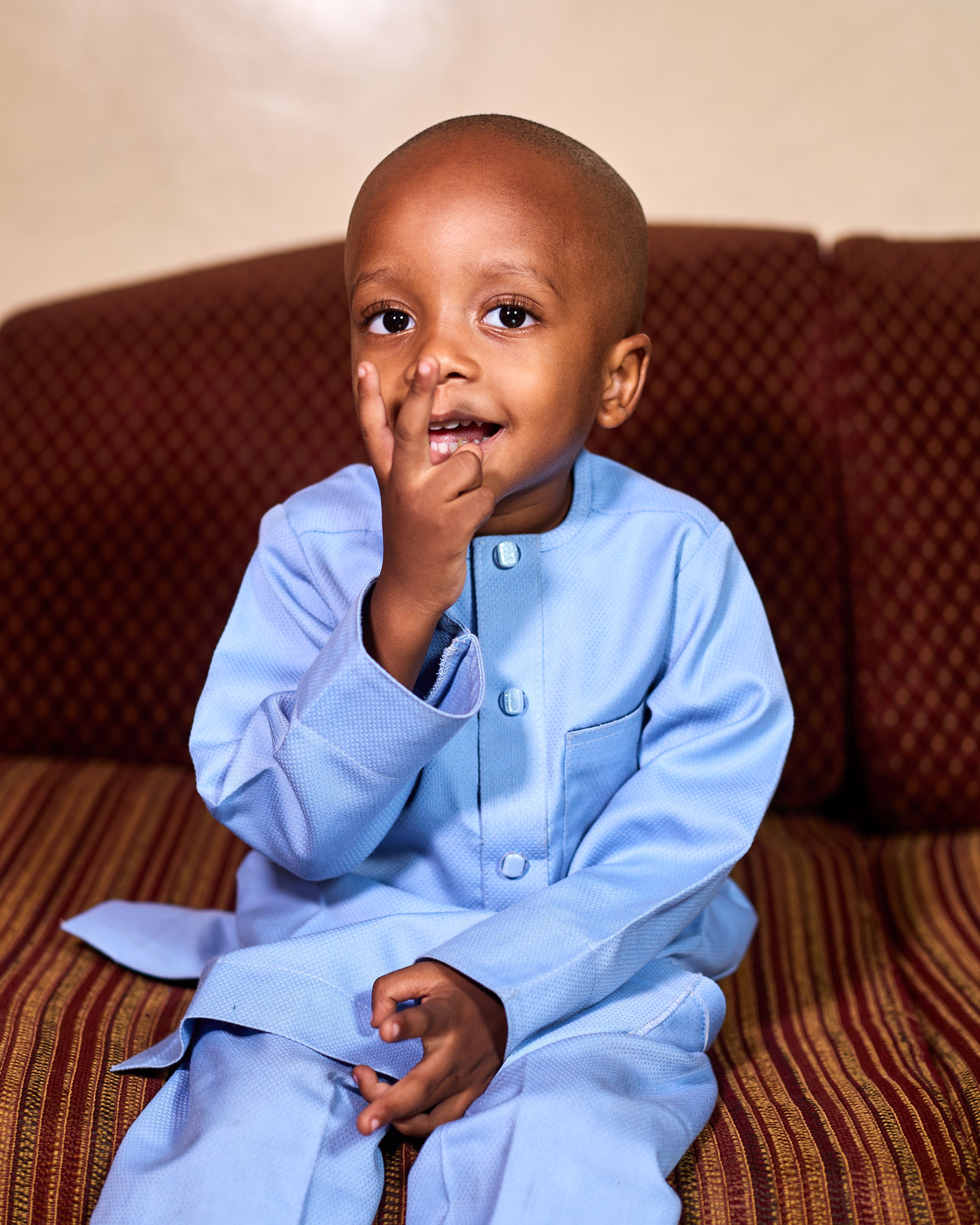 Smiling young boy in blue traditional outfit seated indoors on a sofa in Kaduna, Nigeria.