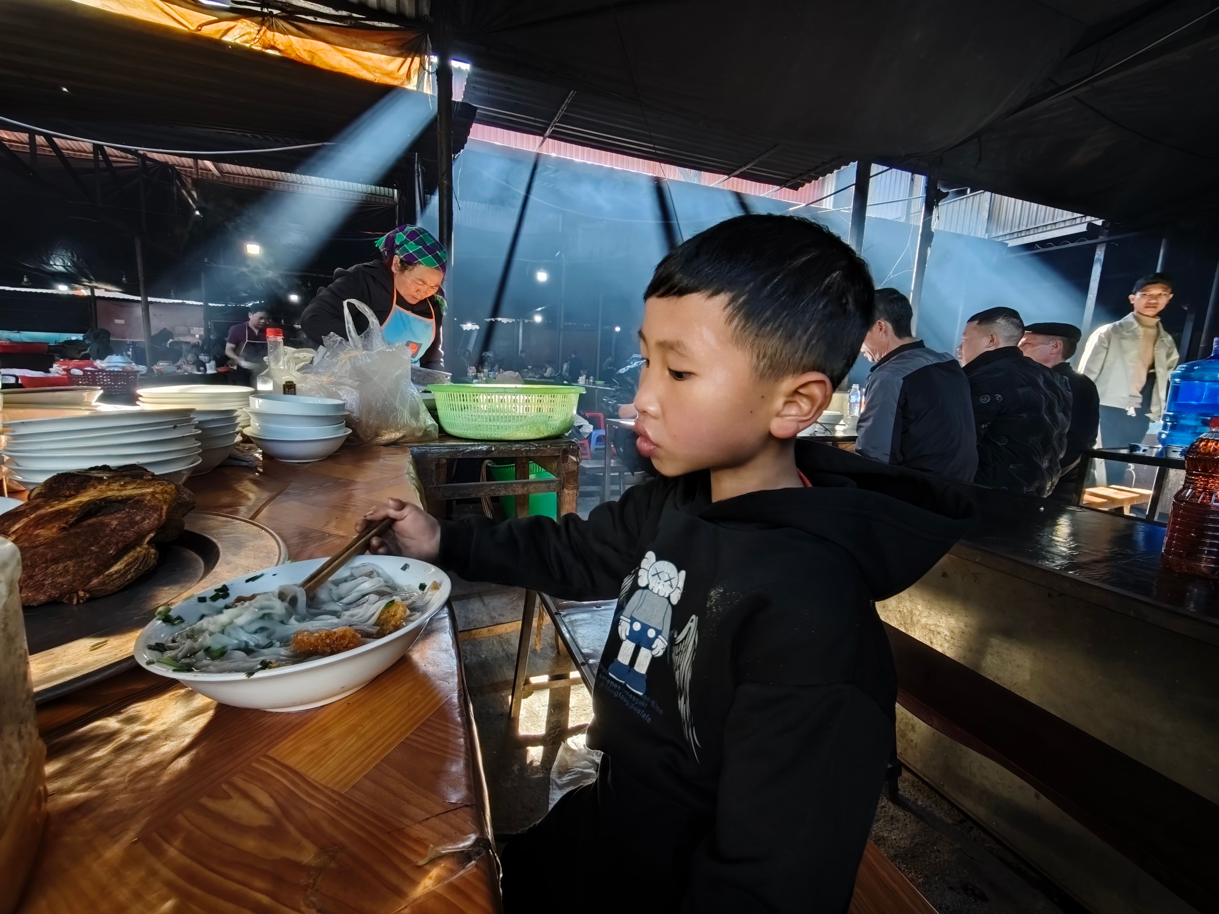 Family with young child at a Singapore hawker centre, toddler in high chair eating a bowl of noodle soup, busy food stall background