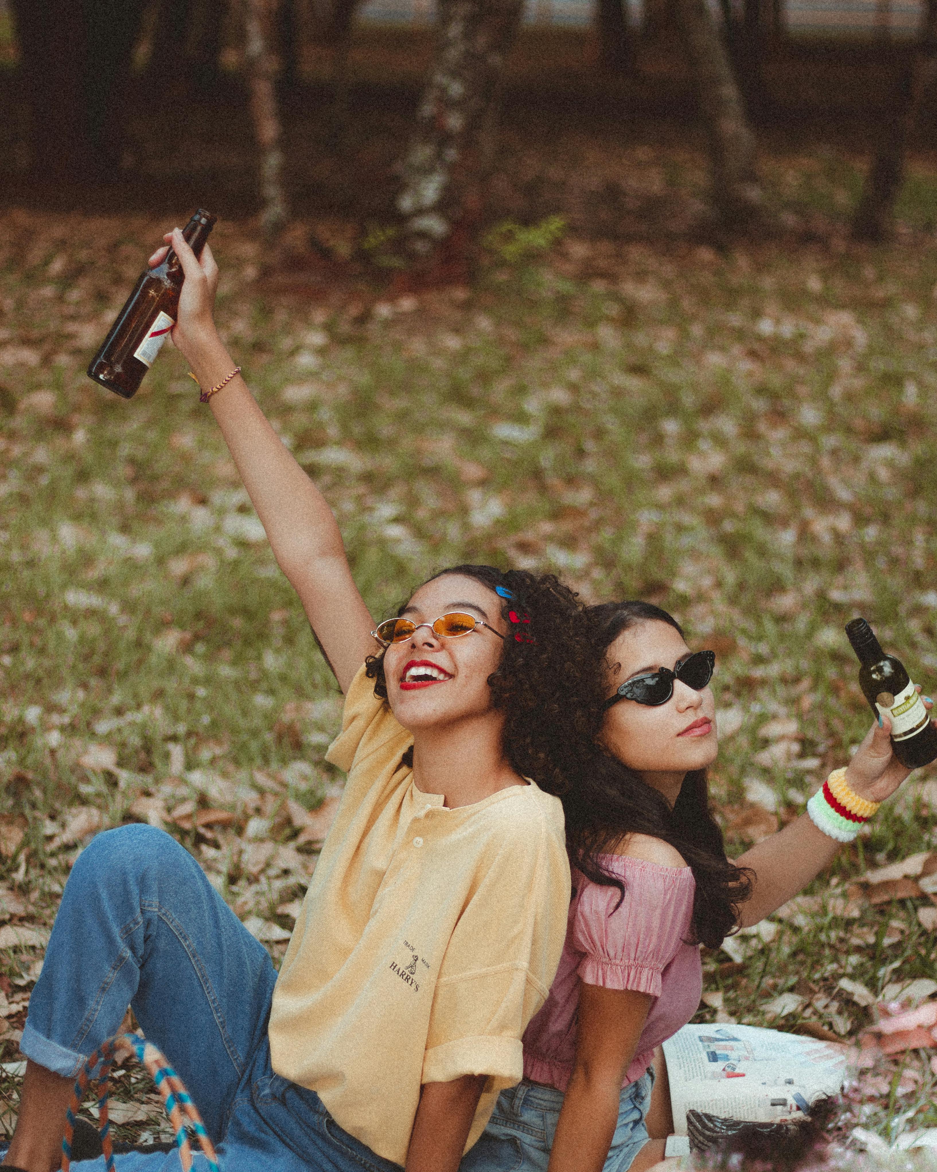 two smiling women holding bottle sitting on green grass field