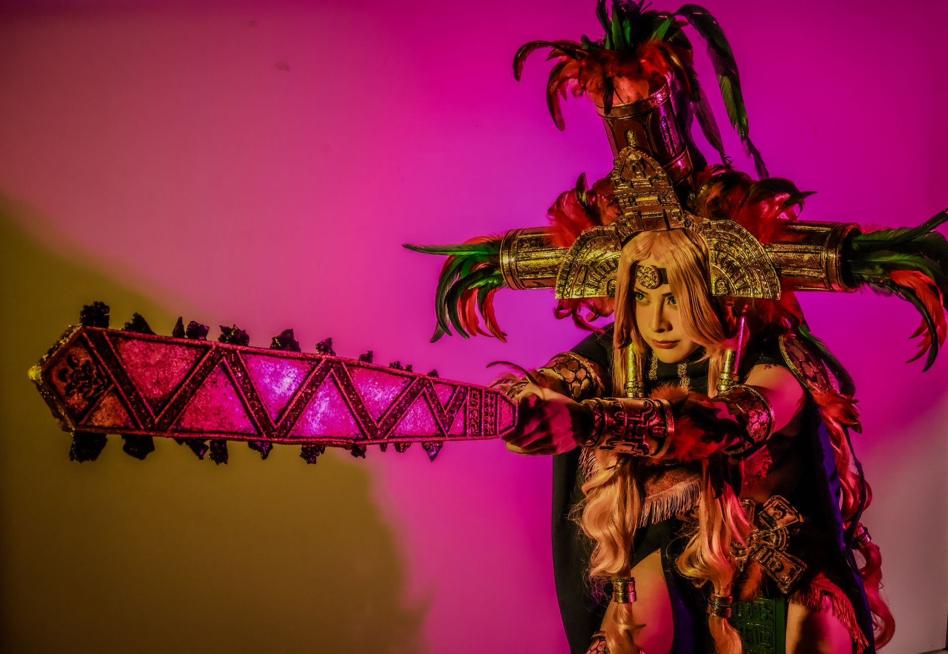 Striking image of a vibrant Aztec warrior cosplay with a feathered headdress and ornate weapon under dramatic lighting.