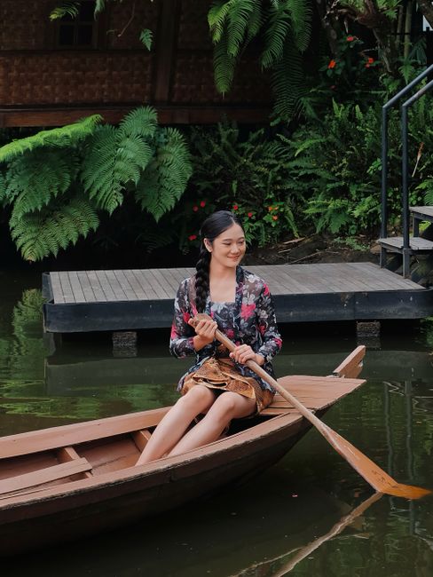 A young woman in a floral dress rows a wooden boat on a tranquil lake in Indonesia.