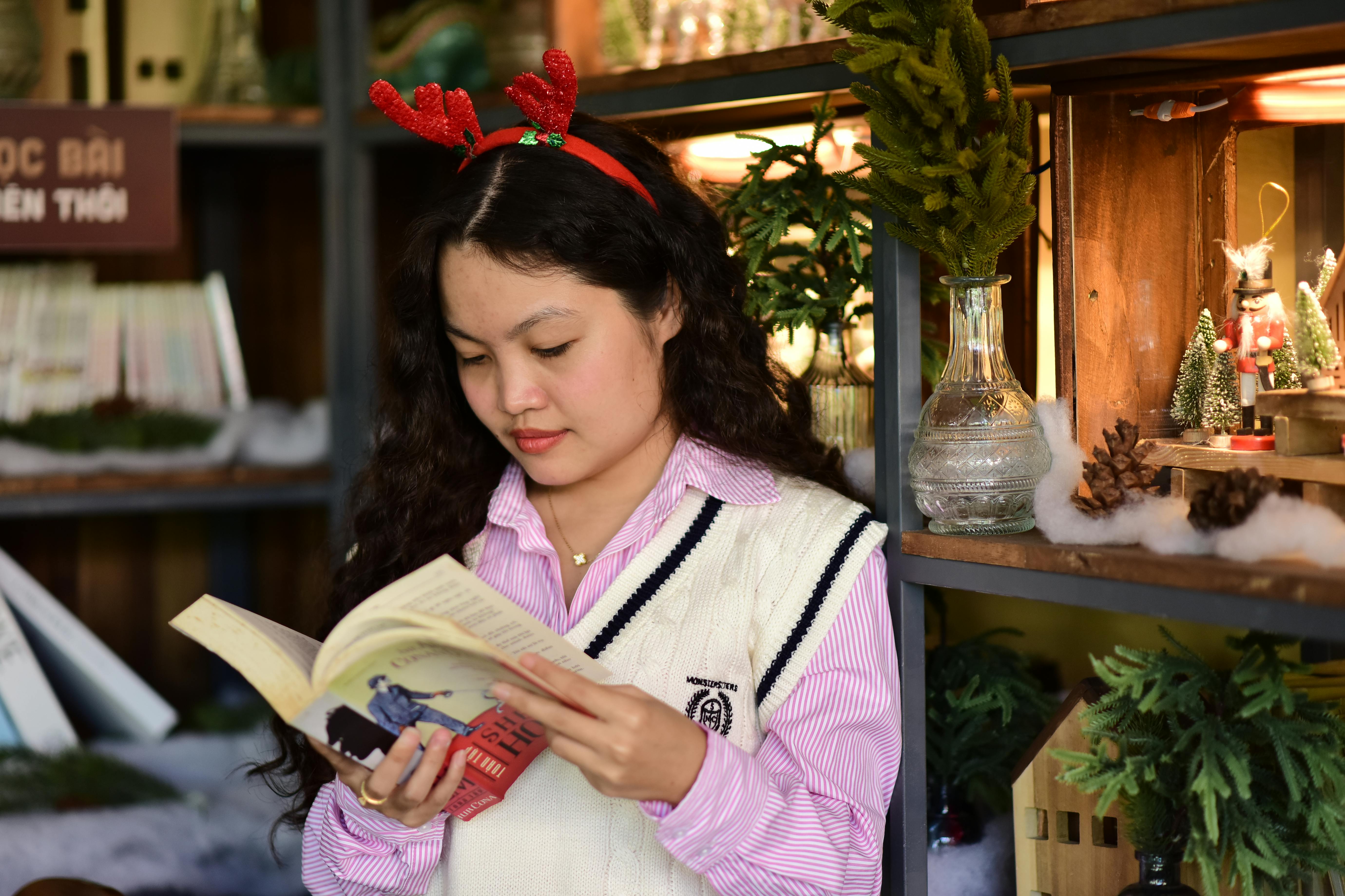 grátis Uma jovem com chifres festivos lê um livro dentro de casa, rodeada por decorações natalinas. Foto profissional