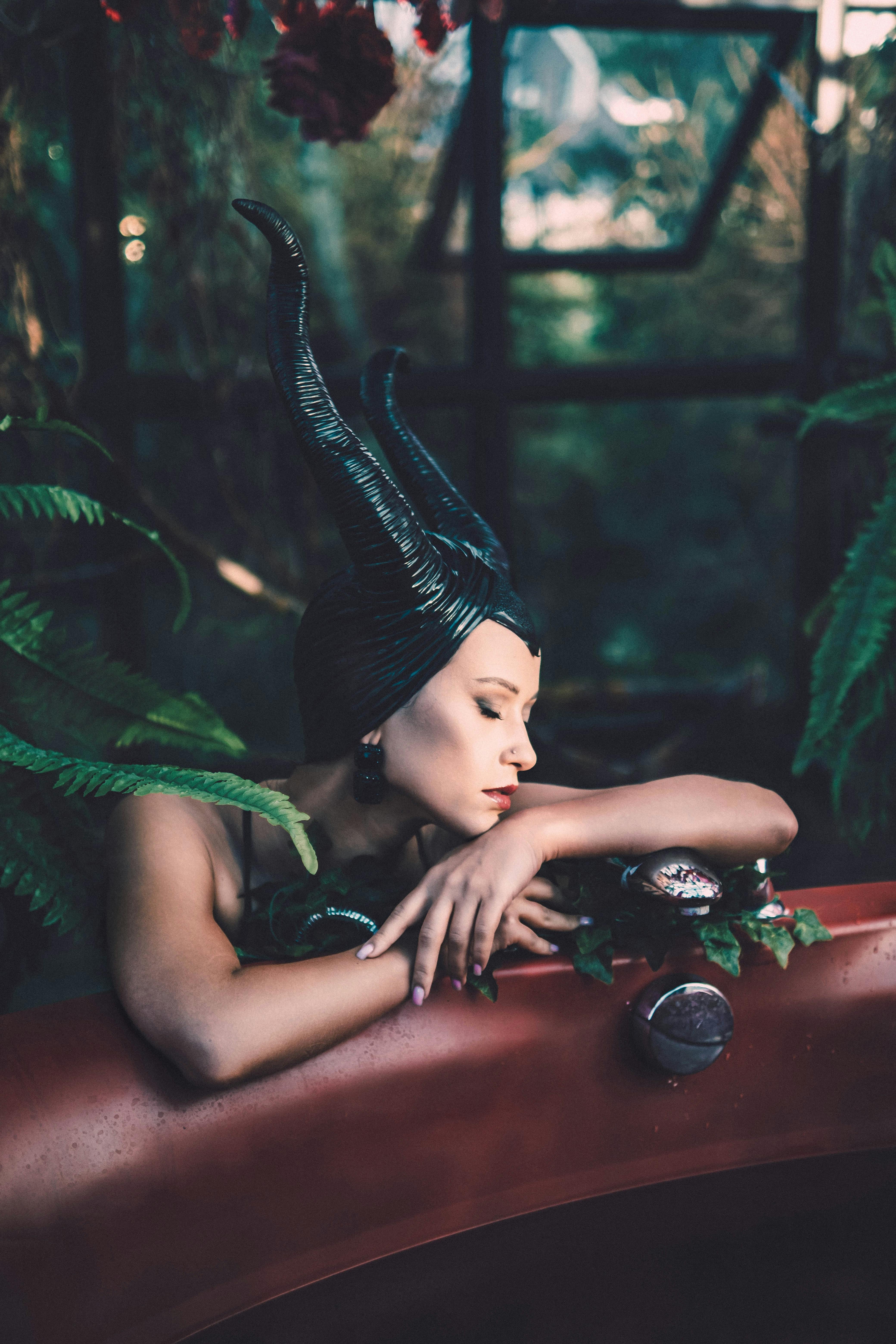grátis Mulher vestida com um traje de fantasia sombria e com chifres relaxando ao lado de uma banheira em meio a uma vegetação exuberante. Foto profissional