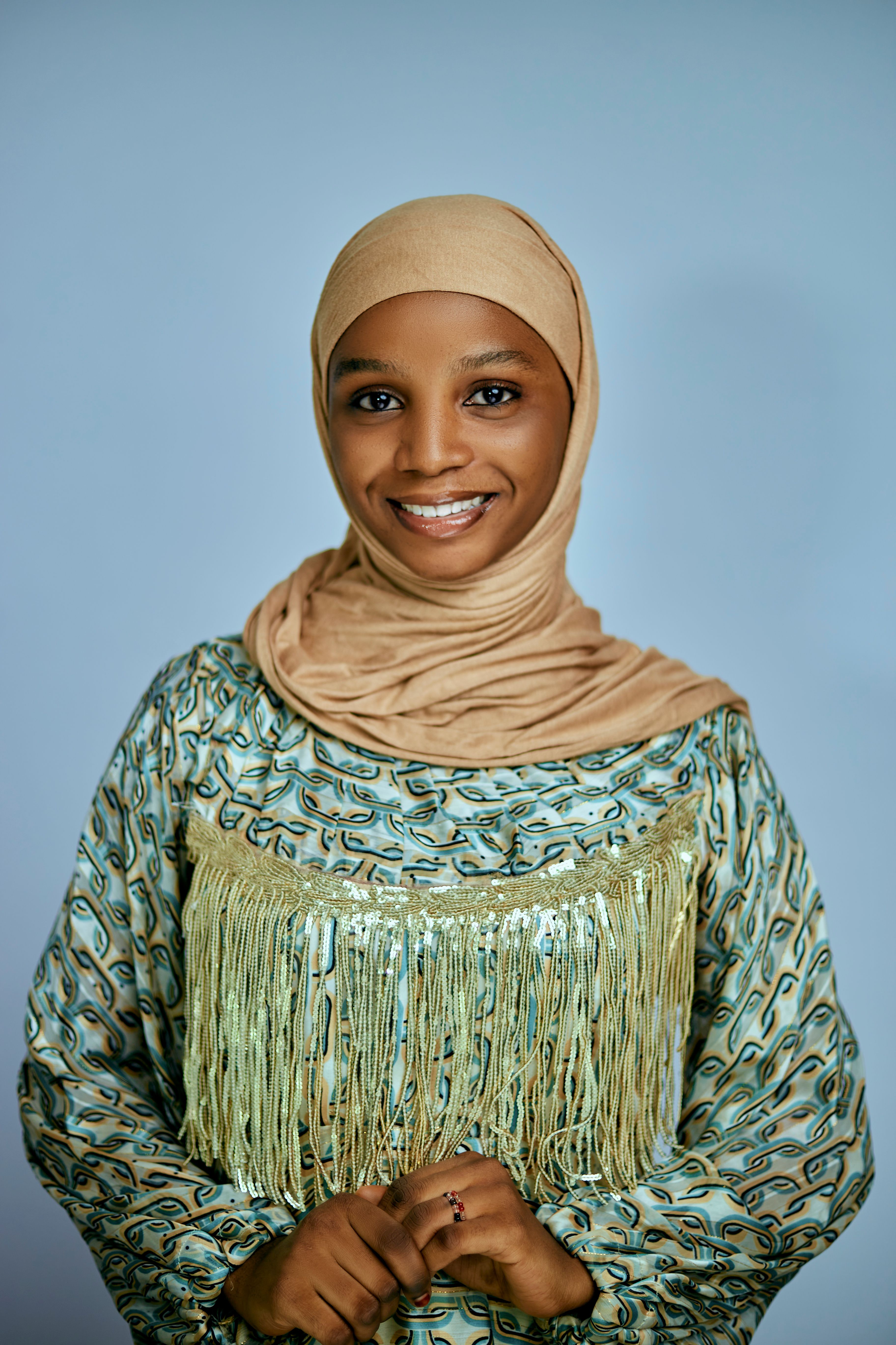A woman in a beige hijab and patterned fringe top smiles softly against a neutral backdrop.