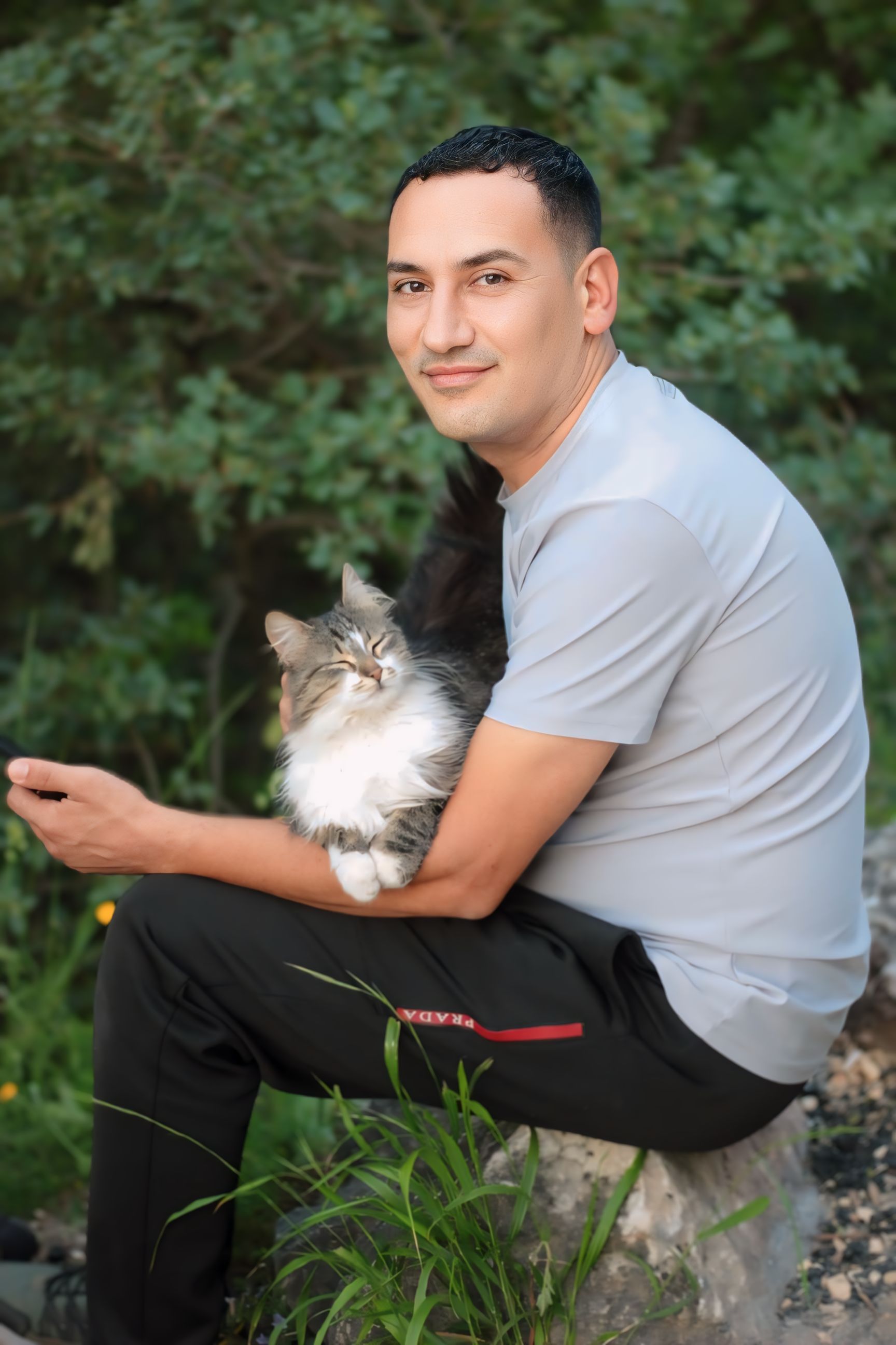 Man sitting outdoors with a fluffy cat on his lap, enjoying a peaceful moment.