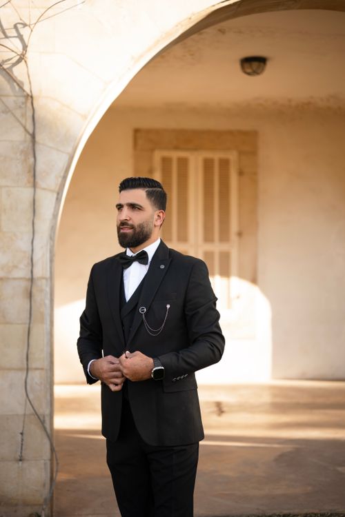 Free Stylishly dressed man in tuxedo poses confidently under sunlight by stone archway. Stock Photo