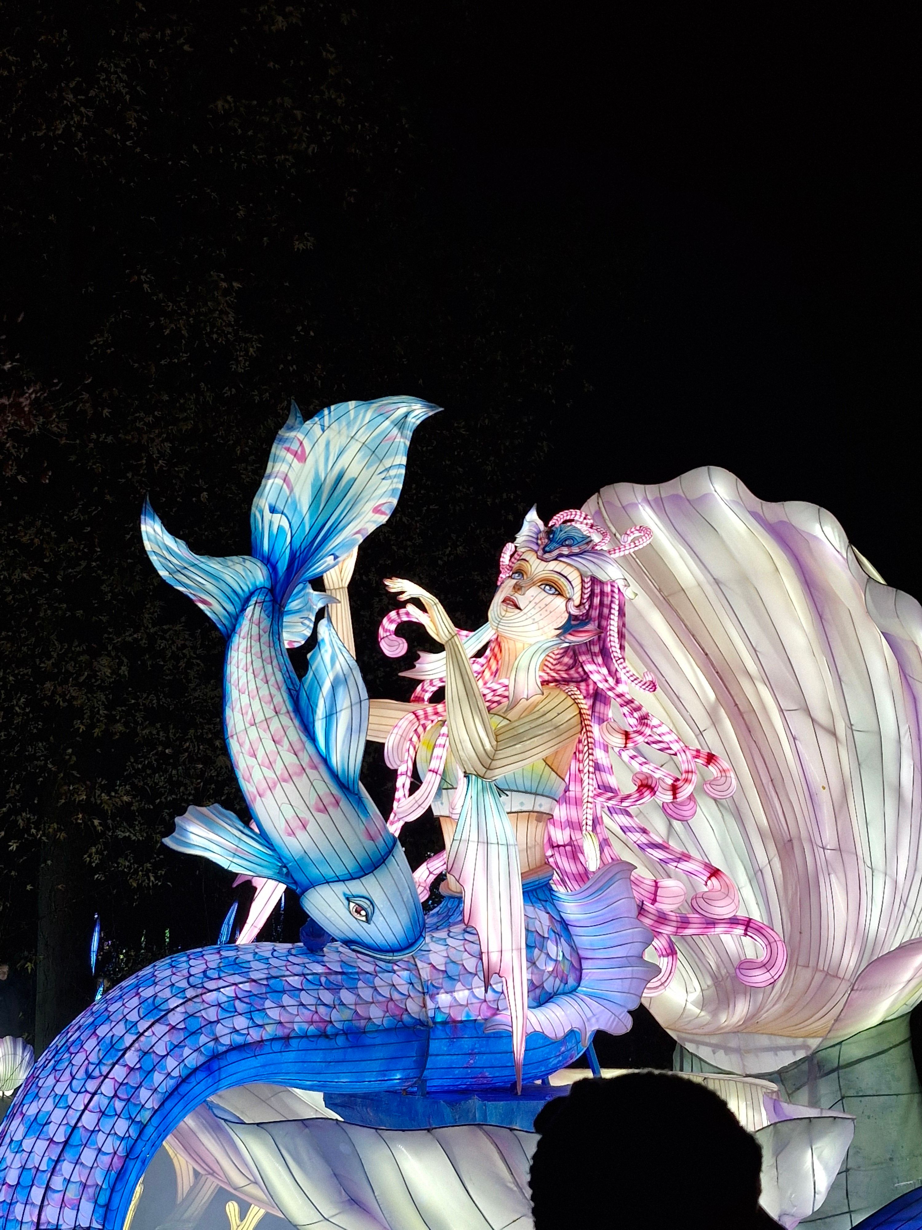 Vibrant mermaid sculpture illuminated against a night sky.