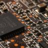 A close-up macro photograph of a modern black microchip surrounded by small electronic components on a printed circuit board.